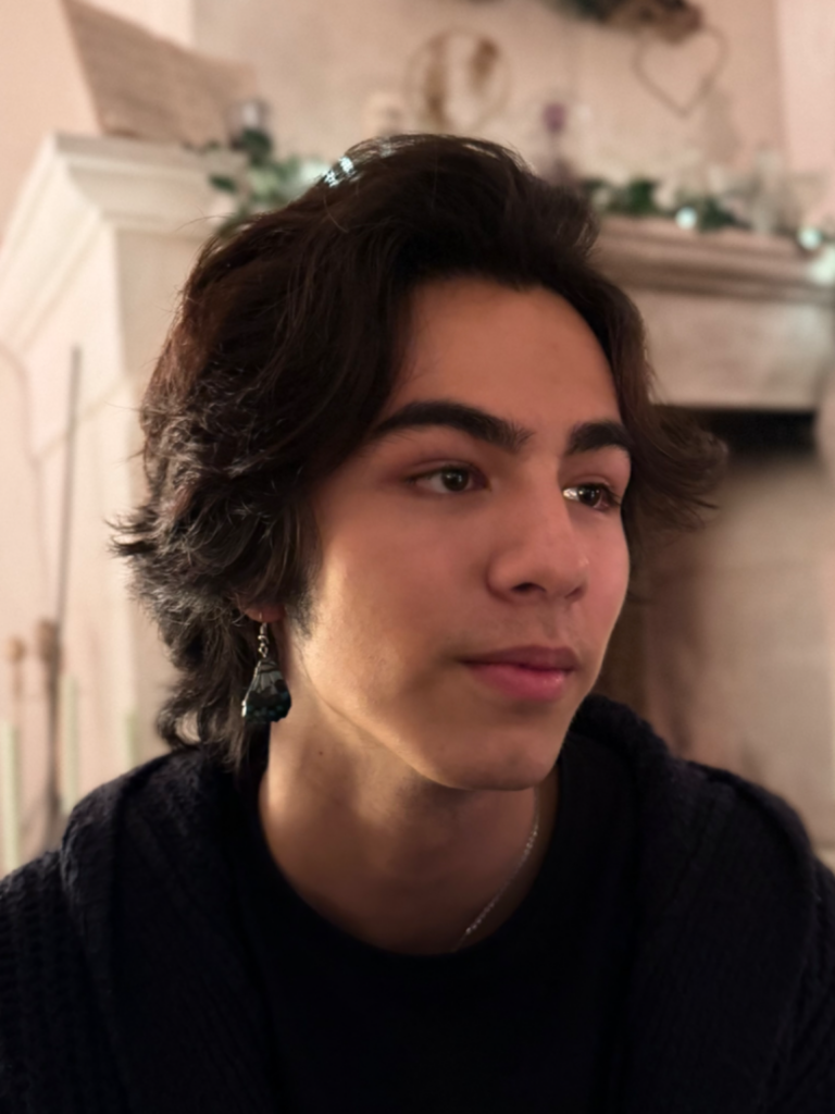 Portrait rapproché d'un jeune homme aux cheveux bruns ondulés, portant un t-shirt noir, un cardigan sombre et une boucle d'oreille pendante foncée. Il regarde pensivement vers la droite.