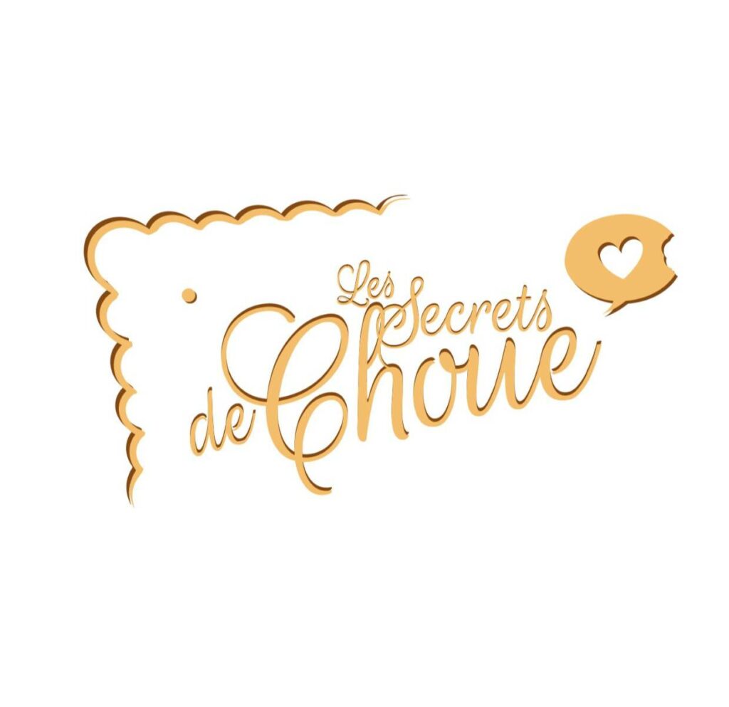 Logo de la marque 'Les Secrets de Choue' sur fond blanc pur. Le texte 'Les Secrets' est écrit en petite police manuscrite marron clair, positionné au-dessus et légèrement à gauche du mot 'Choue', qui est le plus grand et en police cursive épaisse, de couleur caramel doré avec un contour marron foncé. Le coin supérieur gauche est orné d'une bordure ondulée, évoquant un biscuit. En haut à droite, une bulle de dialogue couleur caramel, avec un cœur évidé en son centre et une petite encoche de morsure sur le bord supérieur droit, symbolisant les messages secrets et la gourmandise.