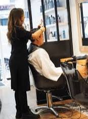 Une coiffeuse professionnelle aux cheveux bruns, vêtue de noir, se tient debout et coiffe méticuleusement les cheveux d'un client assis de dos, portant une cape blanche. La scène se déroule dans un salon de coiffure moderne avec des étagères de produits capillaires visibles en arrière-plan, illustrant l'expertise et le service personnalisé.