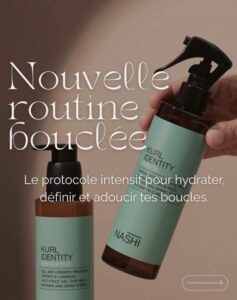 Gros plan détaillé sur deux flacons de produits capillaires Nashi KURL IDENTITY pour cheveux bouclés, sur un fond beige clair. Une main tient un spray volumisant 'kinky fix' (flacon en verre ambré, étiquette vert menthe avec texte blanc, pulvérisateur noir) légèrement incliné vers la droite. En arrière-plan, un flacon pompe de gel anti-frisottis 'bye bye frizz' de la même gamme est visible, partiellement flou. Le texte blanc superposé sur l'image proclame 'Nouvelle routine bouclée' et 'Le protocole intensif pour hydrater, définir et adoucir tes boucles'.