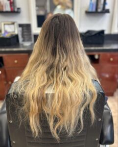 Vue de dos d'une femme aux longs cheveux ondulés, présentant des racines châtain foncé très apparentes sur environ 10-15 cm, contrastant avec un balayage blond doré délavé et légèrement jauni sur les longueurs et pointes. Les cheveux semblent secs, avec des frisottis et des pointes moins définies, reposant sur le dossier noir d'un fauteuil de salon de coiffure. L'arrière-plan est un intérieur de salon flou avec des miroirs et des étagères.