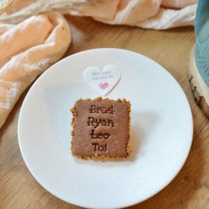 Gros plan, vue de dessus, d'un biscuit artisanal carré au chocolat, de couleur marron clair, avec des bords légèrement croustillants. Le biscuit est gravé en relief avec les noms 'Brad', 'Ryan', 'Leo' et le mot 'Toi', chacun sur une ligne distincte. Il repose au centre d'une assiette en céramique blanche texturée. Au-dessus du biscuit, une petite étiquette en forme de cœur blanc porte le texte 'Mon cœur bat pour toi' et un petit cœur rose. En arrière-plan flou, on distingue un tissu plissé rose pâle parsemé de petits points dorés à gauche, et un morceau de bois clair à droite.