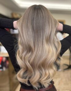 Photo de dos d'une femme aux longs cheveux blonds ondulés et très volumineux. La chevelure présente un balayage multi-tonal, avec des racines châtain clair se fondant en mèches blond cendré et miel vers les pointes. Les cheveux sont brillants, sains, et coiffés en larges boucles souples. Les mains de la femme sont légèrement levées, tenant la masse de cheveux pour en accentuer le volume et le mouvement. Elle porte un haut noir. L'image met en valeur une transformation capillaire réussie.