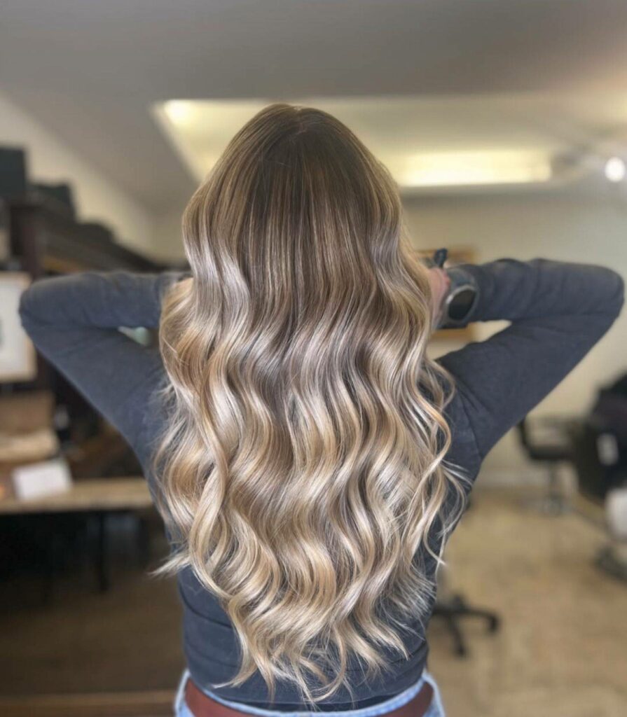 Vue de dos d'une femme aux longs cheveux ondulés, présentant un balayage blond froid et caramel avec des racines châtain clair. Les bras sont levés derrière la tête, mettant en valeur la texture, les reflets brillants et le volume de la chevelure. Le plan est serré sur le dos et les cheveux, dans un intérieur de salon de coiffure avec un arrière-plan flou.