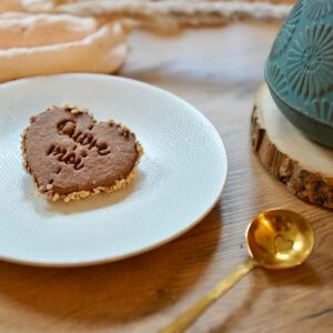 Gros plan sur un biscuit artisanal en forme de cœur couleur chocolat, bordé d'éclats de noisettes, avec le message 'Quoi de neuf ?' en relief, posé sur une assiette blanche texturée. Une cuillère dorée gravée d'un petit cœur est visible au premier plan, tandis qu'une tasse en céramique turquoise ornée de motifs géométriques repose sur un rondin de bois en arrière-plan, le tout sur une table en bois clair.
