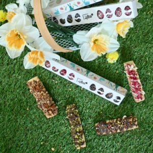 Vue plongeante sur une composition de Pâques avec quatre biscuits artisanaux Les Secrets de Choue aux saveurs variées (pistache chocolat, fruits rouges, noisettes, pépites) disposés sur de l'herbe verte artificielle. Deux coffrets cadeaux blancs à motifs de lapins et œufs, avec des fenêtres en forme d'œufs, sont visibles à côté de narcisses blancs et jaunes et de petits poussins décoratifs. Un panier en bois est partiellement visible en arrière-plan.