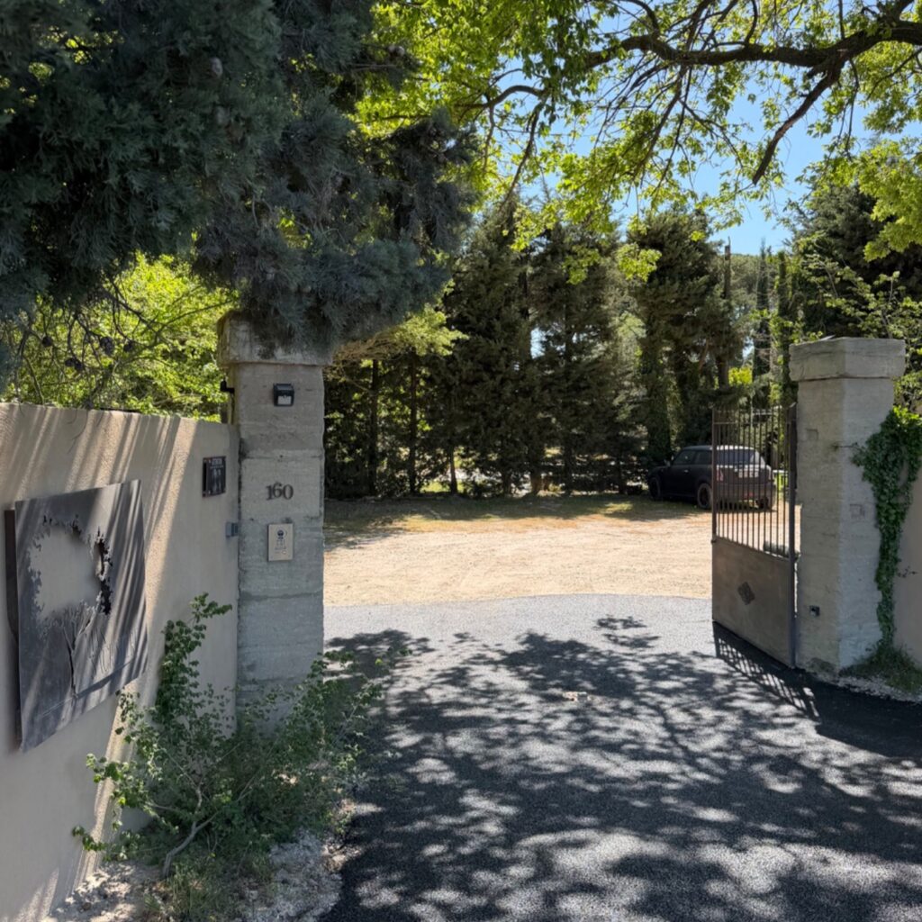 Portail ouvert d'une propriété verdoyante sous le soleil, avec des arbres luxuriants et des ombres sur l'allée, symbolisant l'éveil de la nature et l'arrivée des beaux jours.
