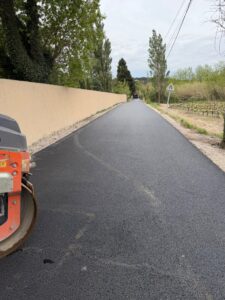 Rouleau compresseur sur une route fraîchement asphaltée, bordée d'un mur clair et de vignes en éveil sous un ciel printanier nuageux.
